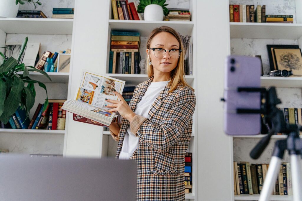 A woman conducting an online class from her home library, using a tripod and smartphone.