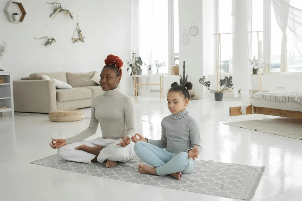 A young African American woman with her daughter sits in meditation in Lotus pose, modeling mindfulness practices.