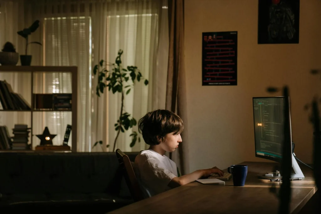 A young boy intensely focused on coding on a computer in a cozy, dimly-lit room.