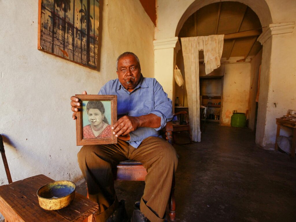 An elderly man seated in a rustic room holds a framed portrait of a woman, showcasing a treasured memory.