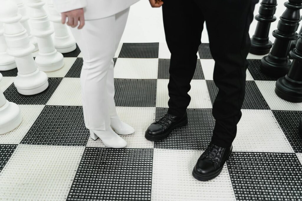 Giant chess pieces on a checkered board with two people in contrasting outfits, symbolizing strategic confrontation.
