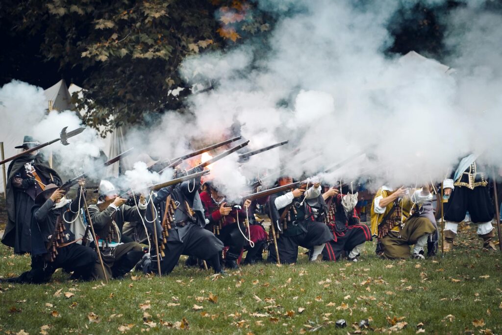 Historical reenactors crouch and fire muskets, with smoke filling the air on a grassy field.