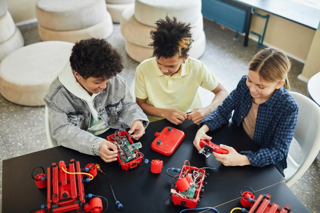 Three children assembling and programming red robotic kits at a table in a bright classroom.