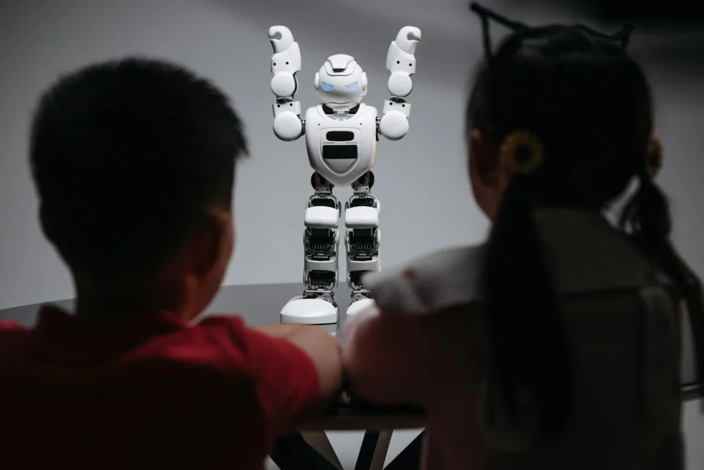 Two children watching a small humanoid robot raise its arms.